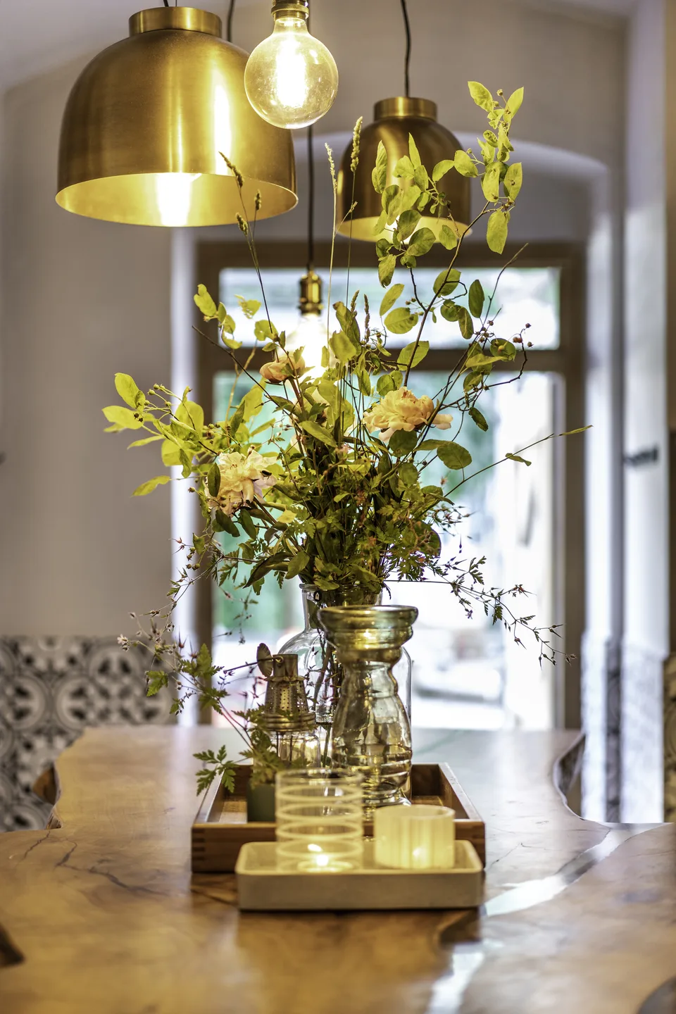 Detailaufnahme eines üppigen Blumengestecks in Glasvase auf dem Naturholztisch des Wirt am Platzl Garsten, darunter Windlichter auf Holztablett, darüber goldene Messing-Pendelleuchten, im Hintergrund Eingangsbereich mit marokkanischen Fliesen.