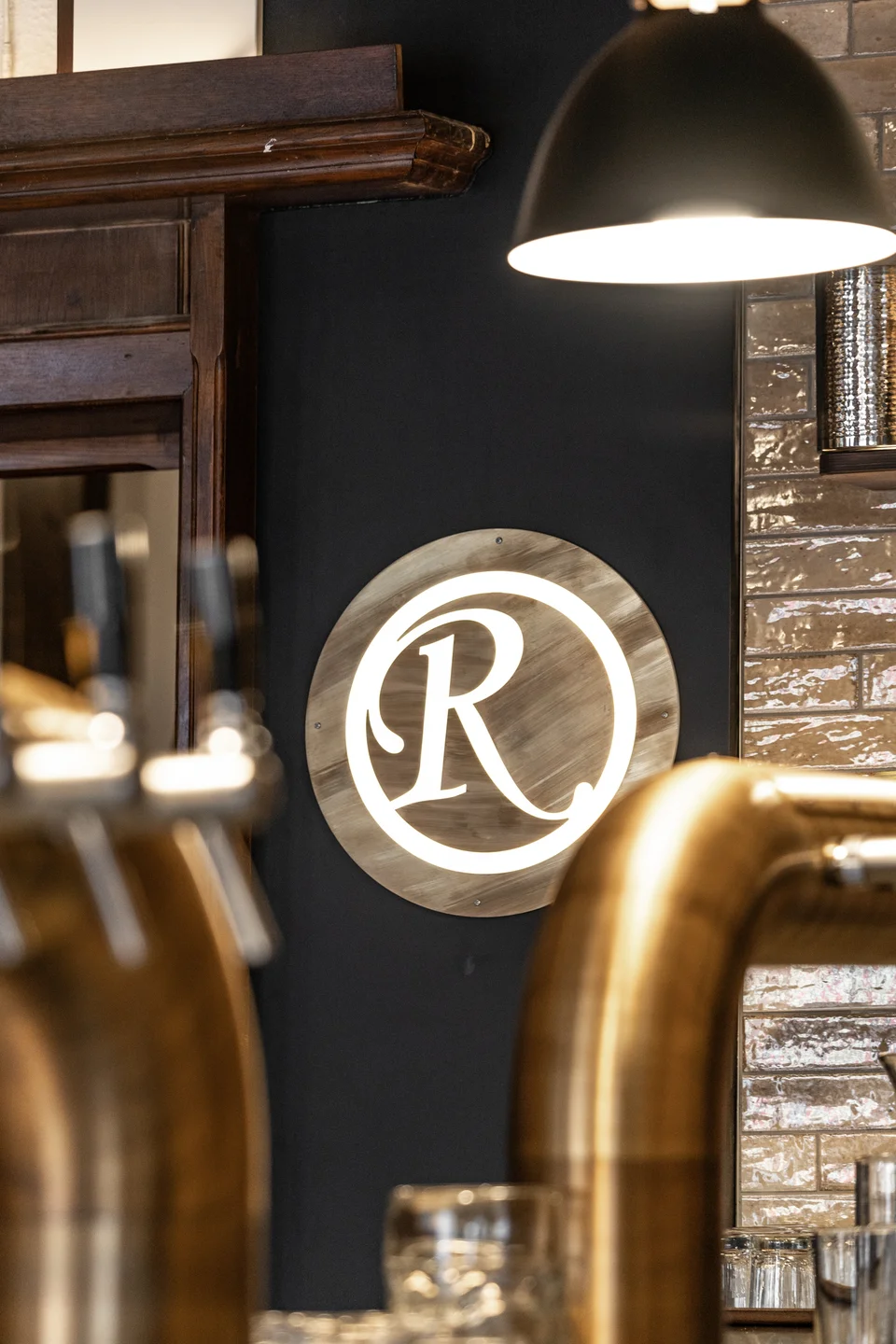 Detailaufnahme des beleuchteten Messinglogos des Restaurant Zum Englischen Reiter in Wien, flankiert von goldener Zapfanlage und schwarzer Pendelleuchte vor glänzender Fliesenwand.