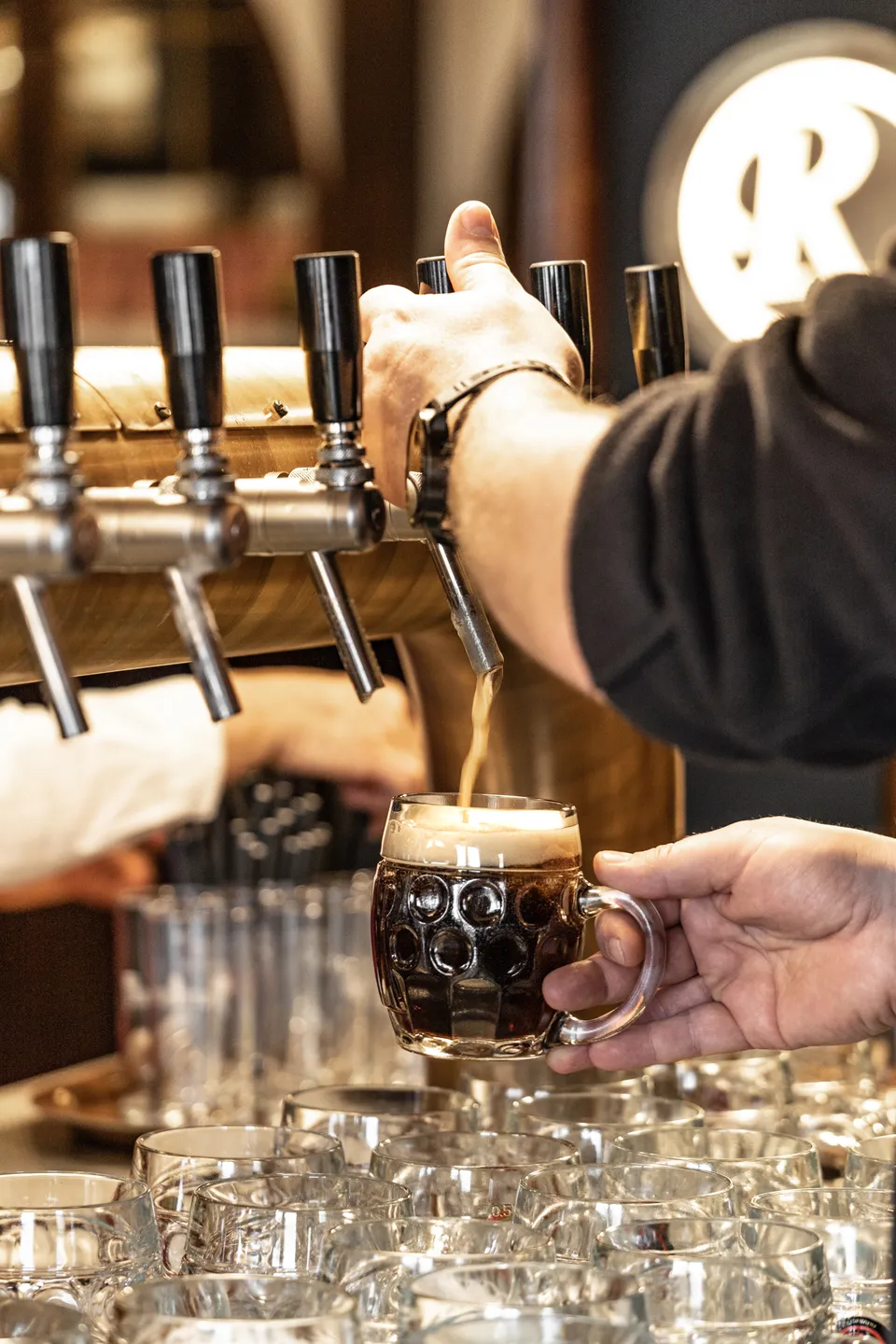 Detailaufnahme beim Bierzapfen im Restaurant Zum Englischen Reiter in Wien – Barkeeper zapft dunkles Bier in einen Bierkrug an der goldenen Zapfanlage, im Hintergrund das beleuchtete Messinglogo.