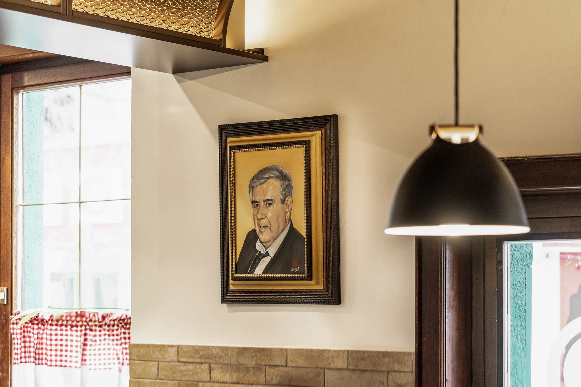 Detailansicht im Restaurant Zum Englischen Reiter Wien mit gerahmtem Portraitgemälde in Goldrahmen an der Wand, schwarzer Pendelleuchte, ornamentaler Messingdecke und rot-weiß karierten Vorhängen am Fenster.