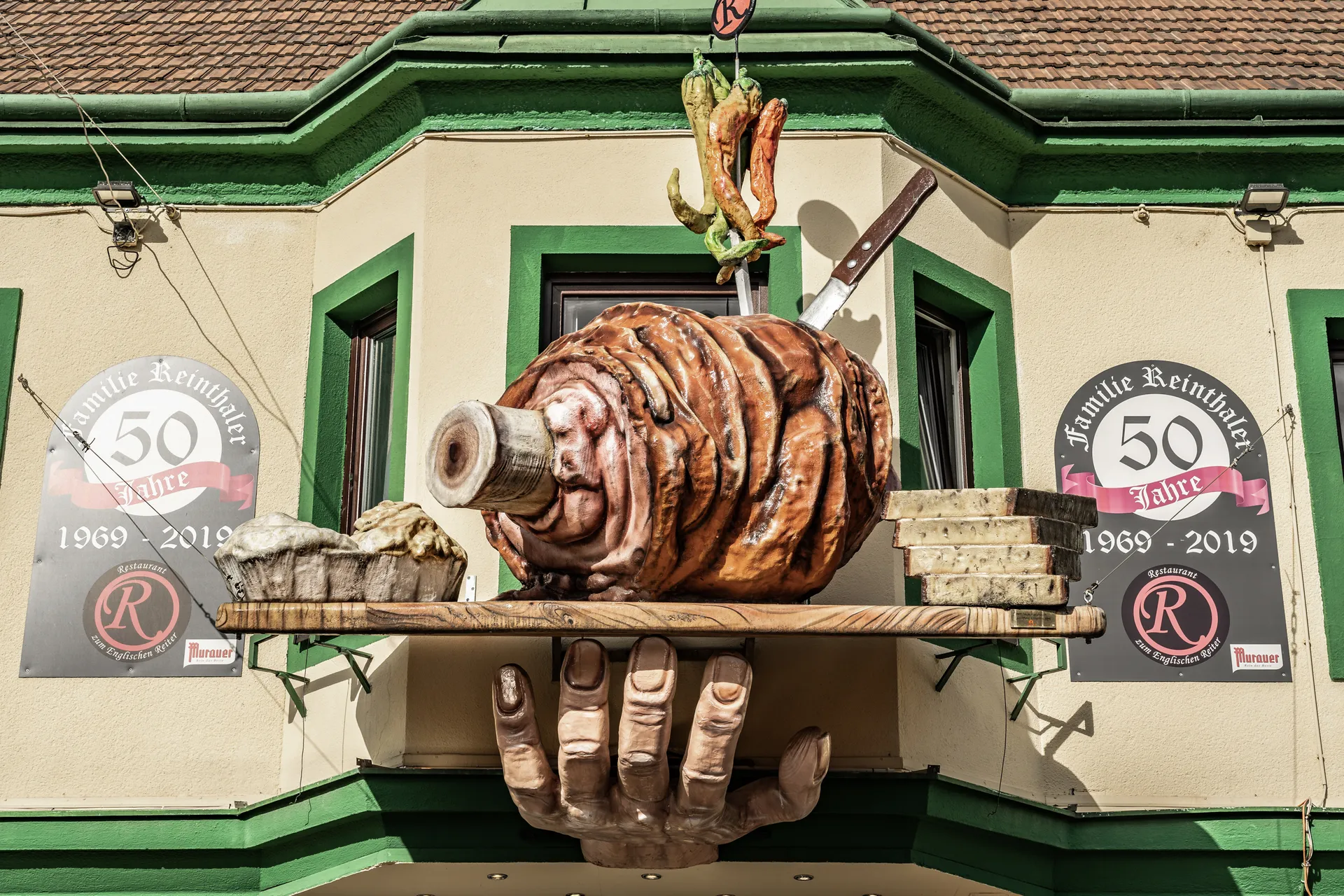 Außenfassade des Restaurant Zum Englischen Reiter in Wien mit monumentaler Stelzenskulptur auf einer Hand, flankiert von zwei Jubiläumsschildern „50 Jahre Familie Reinthaler 1969–2019