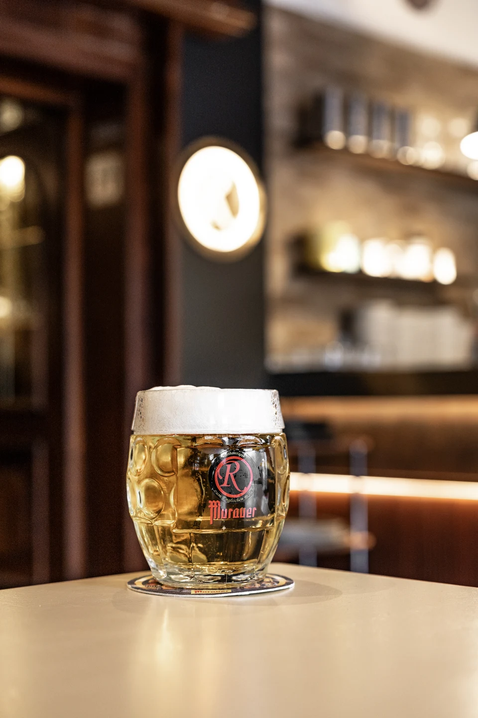 Detailaufnahme eines frisch gezapften Murauer-Bierkrugs mit Logoaufdruck auf dem Tresen des Restaurant Zum Englischen Reiter in Wien, im unscharfen Hintergrund Bar und Glasregale.