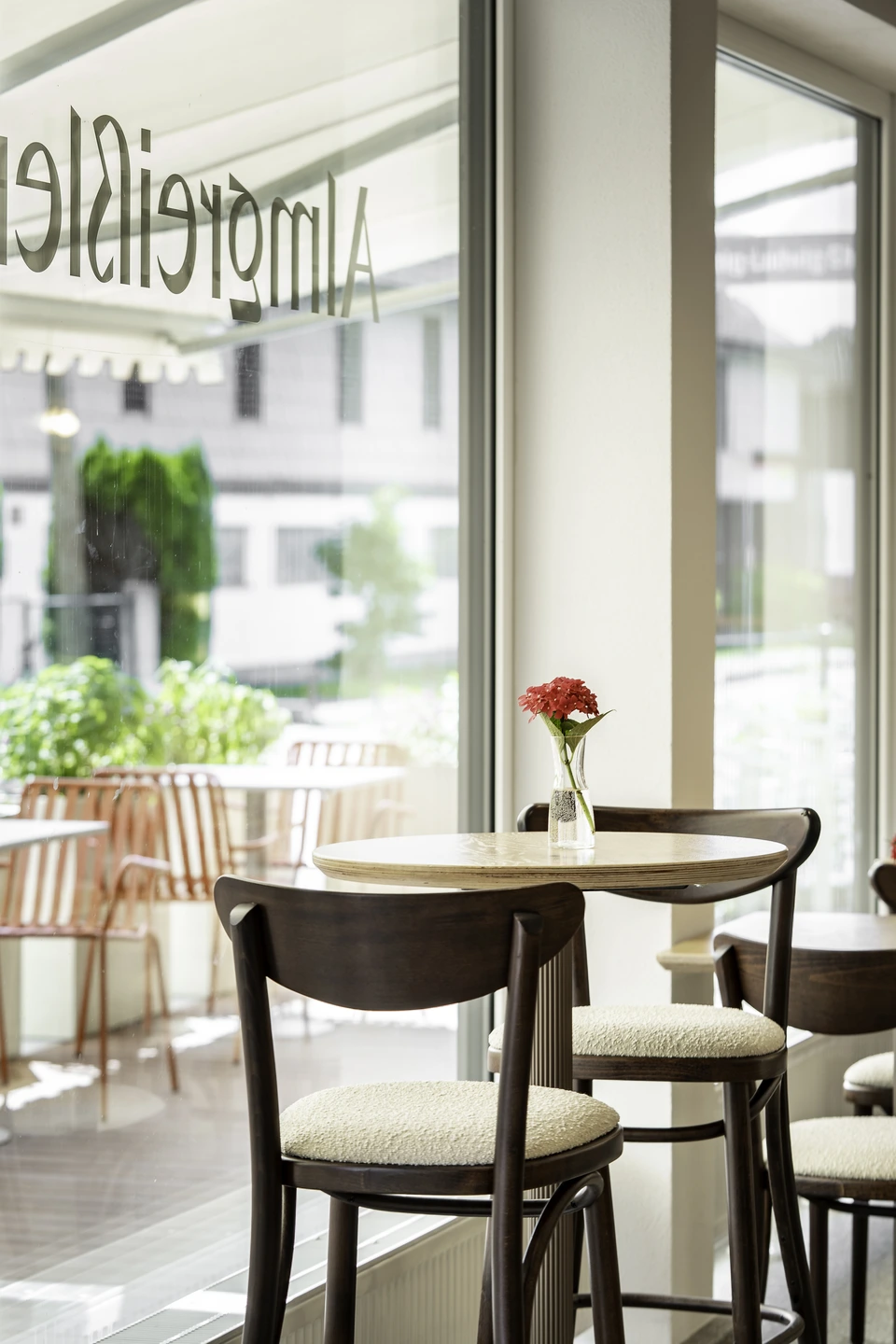 Runder Bistrotisch mit Stühlen und roter Blumenvase vor dem großen Schaufenster der Almgreißlerei Salzburg mit Blick auf die Terrasse.
