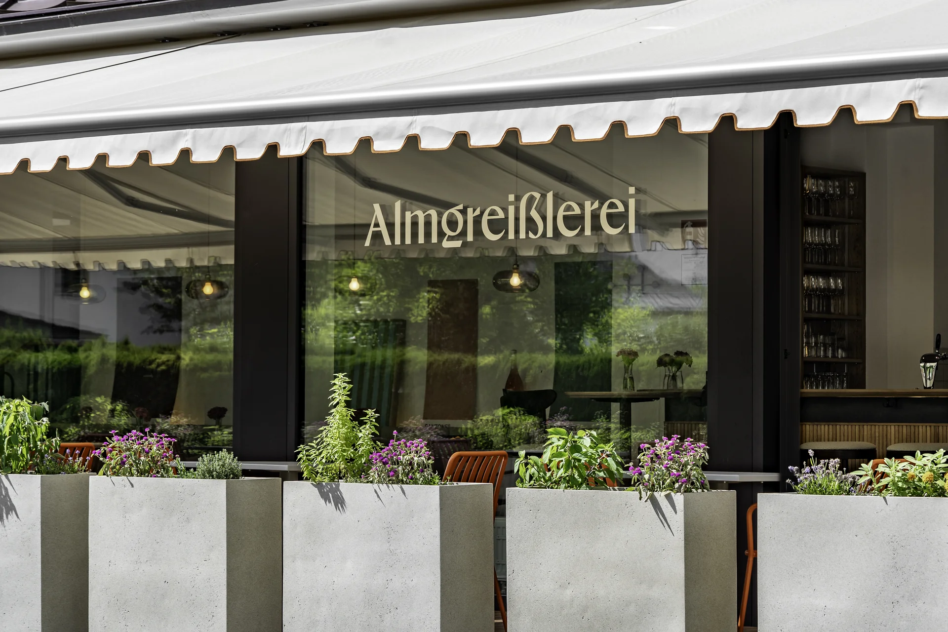 Au&szlig;enansicht der Almgrei&szlig;lerei Salzburg mit Markise, beleuchtetem Schriftzug und bepflanzten Betonk&uuml;beln vor der Terrasse.