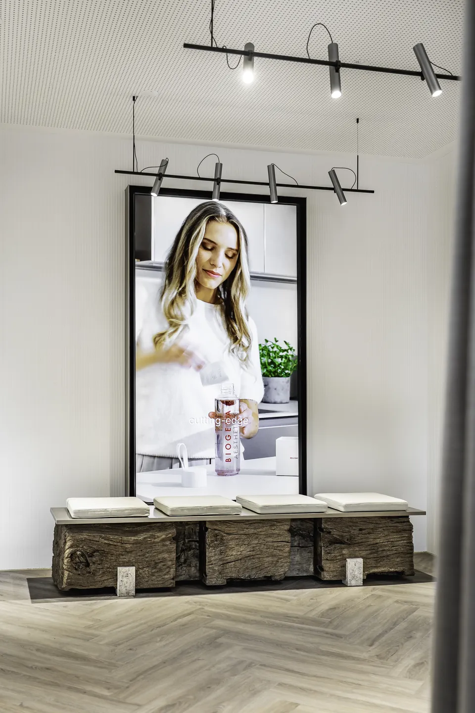 Großformatiger digitaler Screen mit Biogena Aesthetics Werbung über einer rustikalen Holzstammbank mit weißen Kissen in der Biogena Plaza Wien.