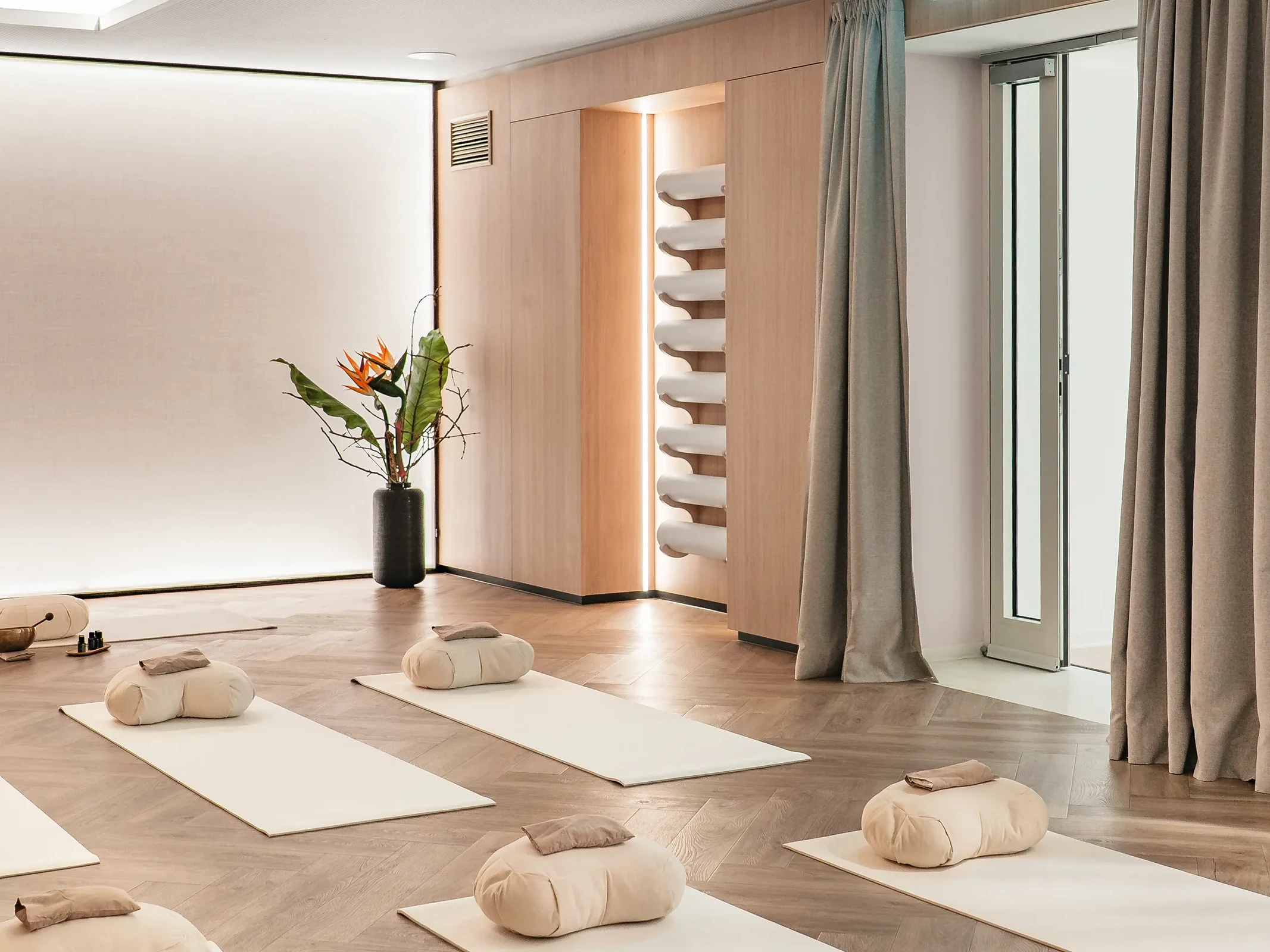 Breath-in Room der Biogena Plaza Wien mit aufgereihten Yogamatten, Meditationskissen, metallischer Wellendecke und Strelitzie vor Holzwand.