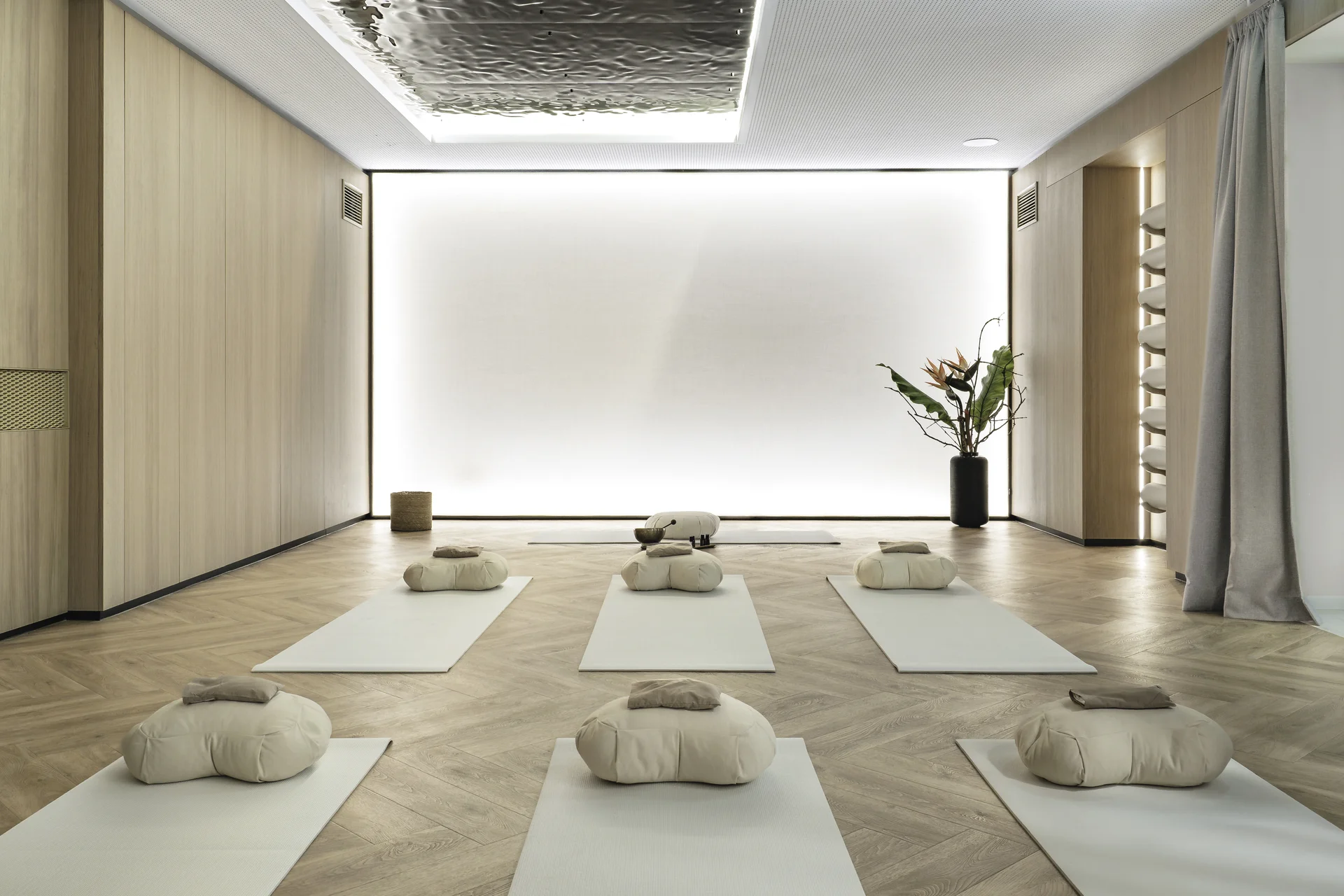 Gesamtansicht des Breath-in Room der Biogena Plaza Wien mit symmetrisch angeordneten Yogamatten und Meditationskissen, hinterleuchteter Lichtwand, organischer Metalldecke und Strelitzie.
