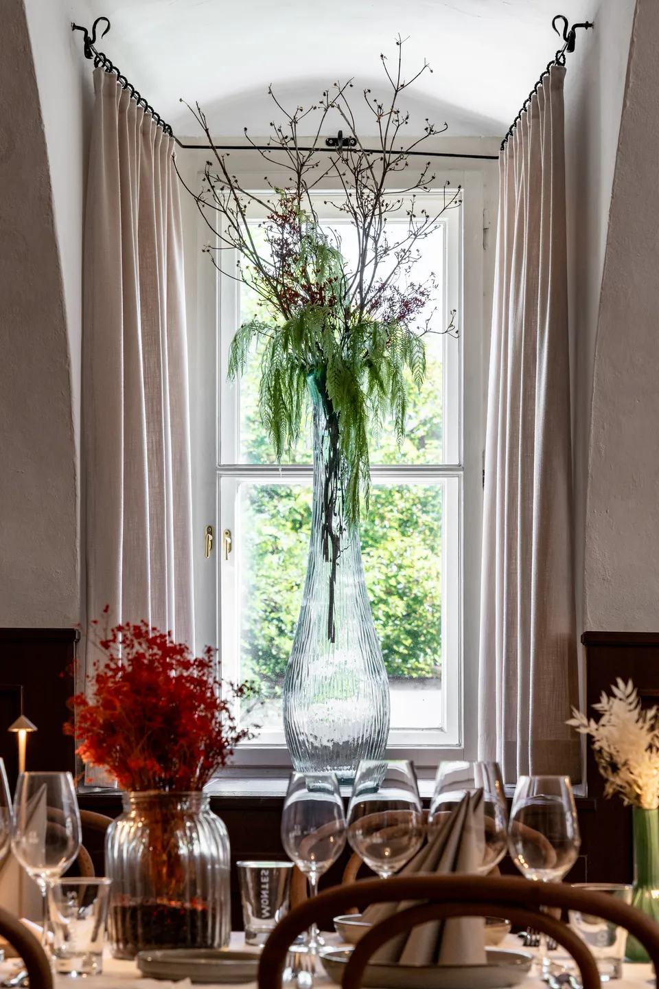 Hohes Fenster im Märchensaal der Fiegnerin mit Leinenvorhängen, großem Glasvasen-Blumenarrangement und festlich gedeckter Tafel davor.