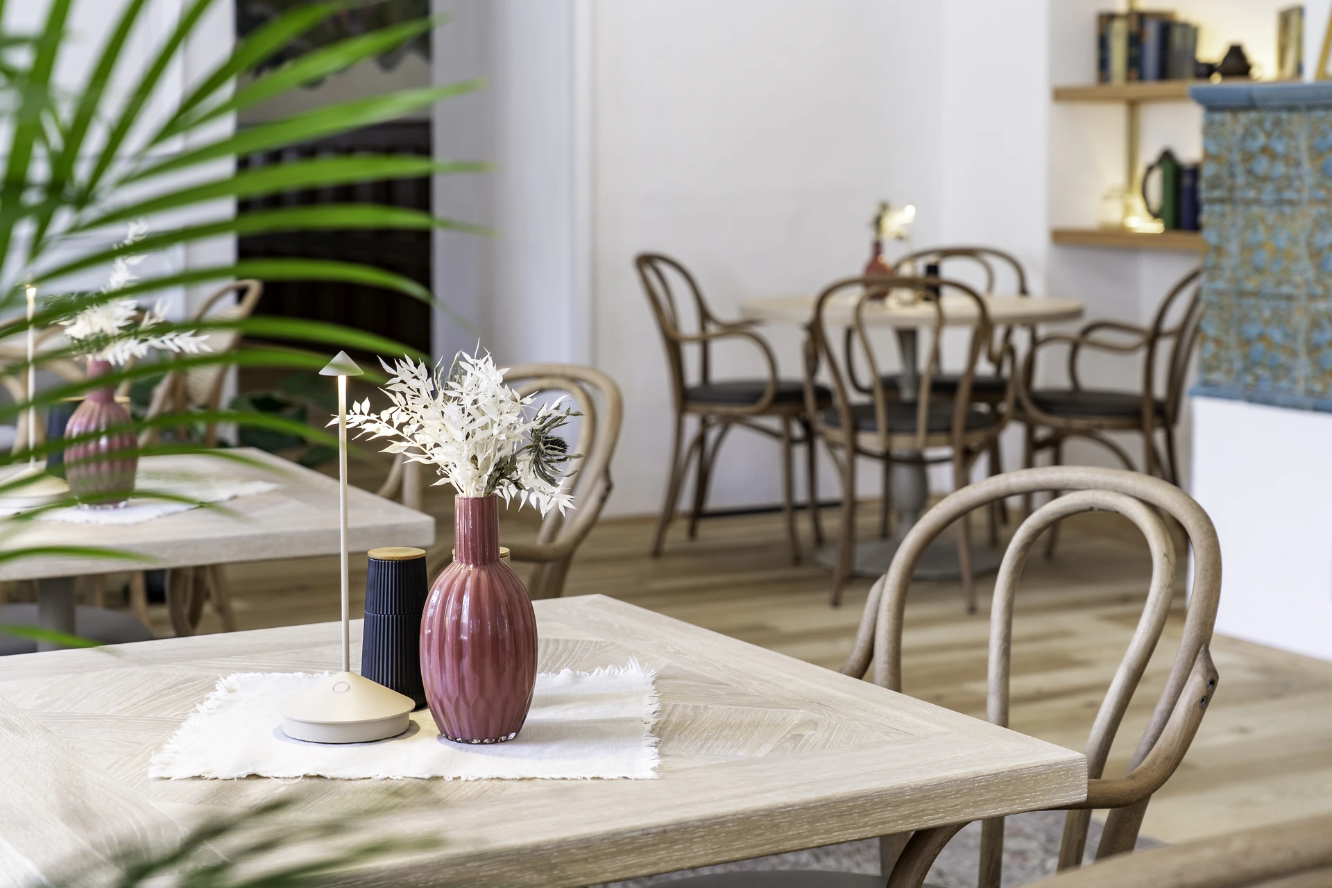 Holztisch in der Fiegnerin im Schloss Fügen mit pinker Keramikvase, weißen Trockenblumen und Akku-Tischleuchte, im Hintergrund Bugholzstühle und Kachelofen.