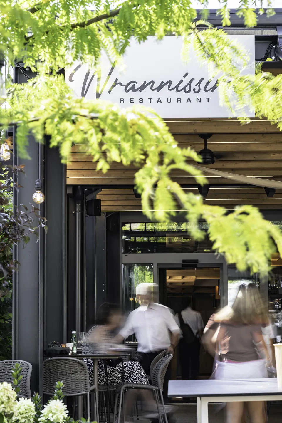 Eingang des Restaurants Wrannissimo in Velden mit Schriftzug über der Tür, Gästen und hellgrünem Blattwerk im Vordergrund.