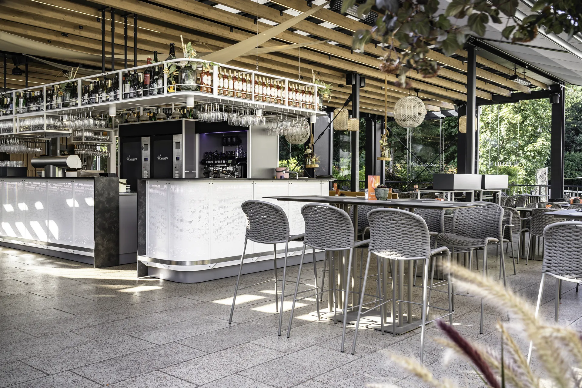Überdachter Gastgarten des Wrannissimo in Velden mit großer Outdoorbar, Holzlamellendecke, Hochtischen, Geflecht-Barhockern und Natursteinboden.