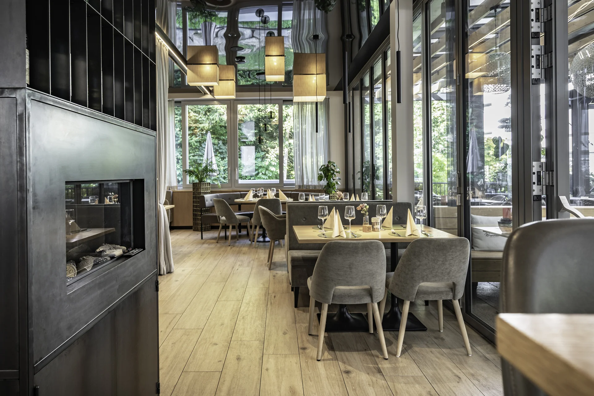 Speisesaal des Wrannissimo in Velden mit schwarzem Kamin, gedeckten Tischen, grauen Polsterstühlen und großer Fensterfront zum Garten.