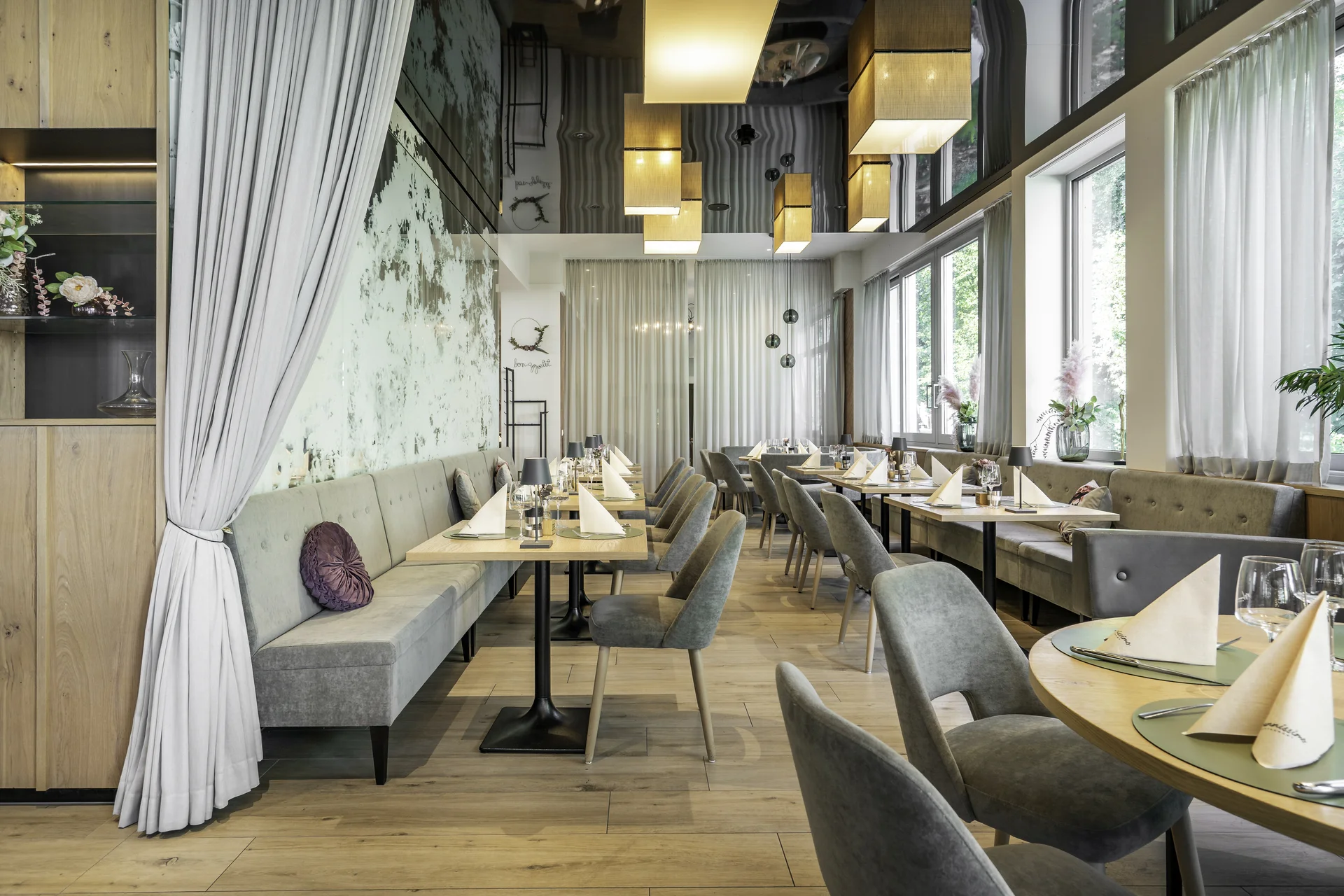 Restaurantbereich des Wrannissimo in Velden mit langer grauer Sitzbank, gedeckten Tischen, hellen Vorhängen, Wandtapete und Pendelleuchten.