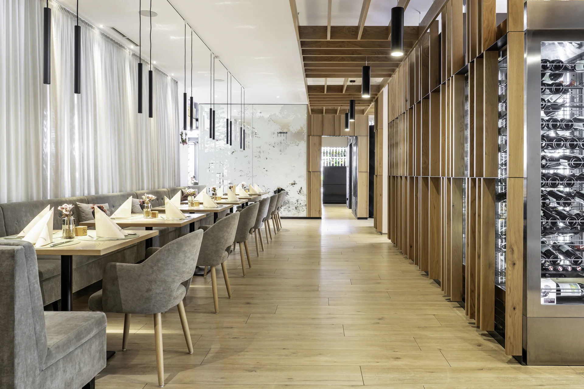 anger Speisesaal des Wrannissimo in Velden mit gepolsterter Sitzbank, gedeckten Tischen, Holzlamellenwand und beleuchtetem Weinklimaschrank.