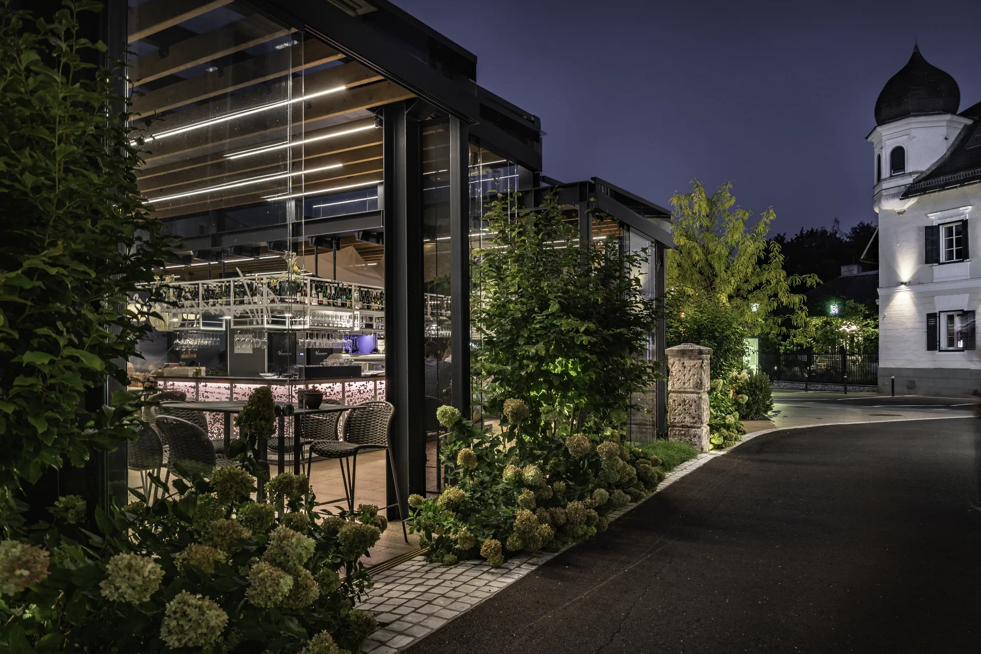 Beleuchteter Gastgarten des Wrannissimo in Velden bei Nacht mit Glasüberdachung, Outdoorbar und Hortensien, im Hintergrund ein Gebäude mit Zwiebelturm.