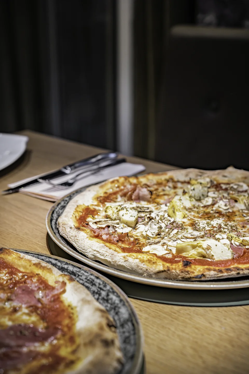 Zwei frisch gebackene Pizzen auf dunklen Keramiktellern auf einem Holztisch im Wrannissimo in Velden.